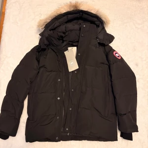Svart dunjacka från Canada Goose - Svart dunjacka från Canada Goose med ikonisk röd logotyp på ärmen. Jackan har en stor huva med beige päls, dragkedja och knappar framtill. Perfekt för kalla vinterdagar och har en clean, klassisk look som funkar till allt.