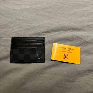 Svart plånbok - Snygg och stilren svart plånbok med rutigt mönster och klassisk logga i hörnet. Perfekt för kort och sedlar, smidig att ha med sig överallt. Designen är minimalistisk och passar dig som gillar exklusiva accessoarer.