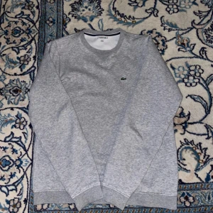 Grå sweatshirt från Lacoste - Grå sweatshirt från Lacoste  med klassisk rund hals och långärmad passform. Tröjan har det ikoniska gröna krokodilmärket broderat på bröstet. Mjuk och skön bomullsmix, perfekt för chill dagar. Ribbstickade muddar vid ärmslut och nederkant.