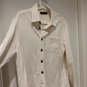  skjorta/overshirt från Morris - Cream/vit skjorta från Morris Stockholm i storlek M. Skjortan har klassisk krage, bröstficka och svarta knappar framtill. Materialet är mjukt och känns som bomull, perfekt för en clean och stilren look. Passformen är normal och ärmarna är långärmade.