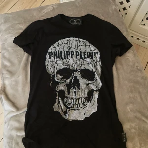 Svart t-shirt med döskalle från Philipp Plein - Svart t-shirt från Philipp Plein Platinum Cut med stor silverfärgad döskalle framtill och logotyp över trycket. T-shirten har korta ärmar och rund hals. Materialet är mjuk bomull och passformen är normal. Perfekt för dig som gillar edgy och exklusiv streetwear.