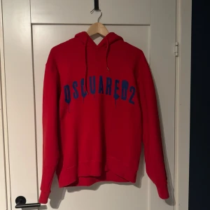 Röd DSQUARED2 hoodie med blått tryck - Säljer en röd hoodie från DSQUARED2 i storlek M med blått tryck på bröstet. Tröjan har huva med snörning och långa ärmar. Materialet är mjuk bomull och passformen är normal. Perfekt för dig som gillar streetwear och snygga loggor.