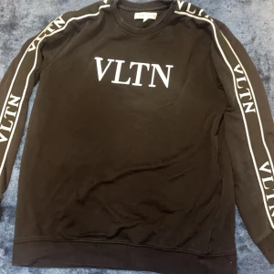 Svart Valentino sweatshirt med VLTN-logga - Svart sweatshirt från Valentino med stor vit VLTN-logga på bröstet och vita VLTN-detaljer längs båda ärmarna. Tröjan har rund halsringning och är långärmad, perfekt för en clean och trendig streetstyle-look. Storlek L men passar M