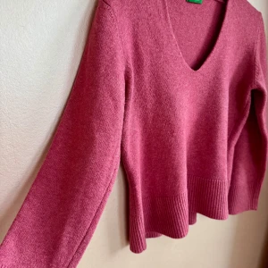 Rosa stickad tröja från Benetton - Rosa stickad tröja från United Colors of Benetton med v-ringning och bred ribbad kant nertill. Långärmad modell i ull, perfekt för kyliga dagar.