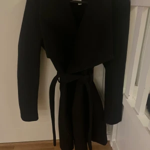 Svart kappa med knytning runt midja från Vila - Snygg svart kappa från Vila med bred krage och knytbälte i midjan. Den är knappt använd. Kappan är lagom tjock och passar perfekt till höst vår och vinter. 