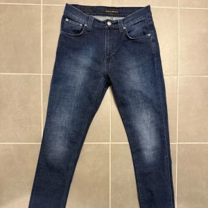 Mörkblå jeans från Nudie Jeans Co - Snygga mörkblå jeans från Nudie Jeans Co med klassisk femficksdesign och subtila slitningar. Jeansen har raka ben, normal passform och är tillverkade i mjukt bomullsmaterial. Bakfickorna har Nudie Jeans signaturbroderi och en svart läderpatch i midjan. Storlek 30x32