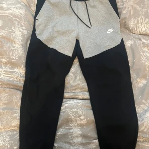 Nike svart-grå mjukisbyxor med snörning - Nike mjukisbyxor i svart och ljusgrått med snörning i midjan och vit Nike-logga på vänster sida. Byxorna har en sportig look, dragkedjeficka bak och är tillverkade i mjukt material för maximal komfort. I ny skick.