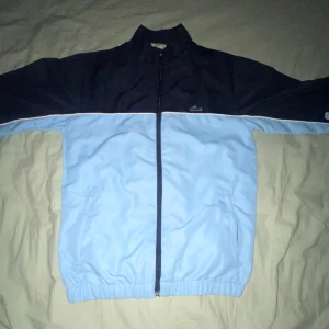 Blå och svart vindjacka från Lacoste - Säljer en snygg vindjacka från Lacoste i ljusblått och svart med vit piping. Jackan har dragkedja framtill, ståkrage och elastisk nederkant. Stor Lacoste-logga på ryggen och liten krokodillogo på bröstet. Perfekt för dig som gillar sportig stil.