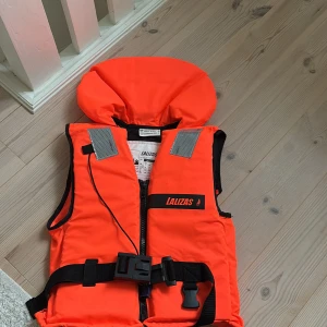 Orange flytväst från Lalizas - Säljer en orange flytväst från Lalizas med svart bälte och spänne framtill. Västen har reflexdetaljer på axlarna och en hög krage för extra säkerhet. Passar vuxna med bröststorlek 80-90 cm och vikt 40-50 kg. Perfekt för båtliv och vattenaktiviteter.