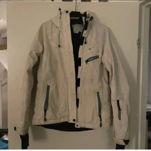 Beige vindjacka med huva från Peak - Snygg vit vindjacka med huva och flera praktiska fickor med gröna dragkedjor. Jackan har lång ärm med justerbara muddar, snörning vid huvan och coola svarta detaljer på framsidan. Perfekt för blåsiga dagar och outdoor-aktiviteter.