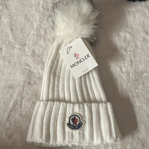 Vit Moncler mössa med pälsboll - Ribbstickad vit mössa från Moncler med stor fluffig pälsboll i vitt och svarta toppar. Klassisk Moncler-logga framtill. Perfekt accessoar för kalla dagar med exklusiv känsla och snygg design.