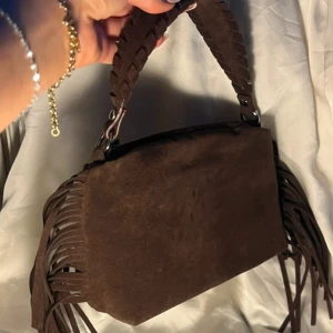 Brun mocka handväska med fransar - Snygg brun handväska i mocka med flätat handtag och långa fransar på sidorna. Väskan har en rektangulär form och cool western-vibe. Perfekt för dig som vill sticka ut med detaljer och mjukt material.