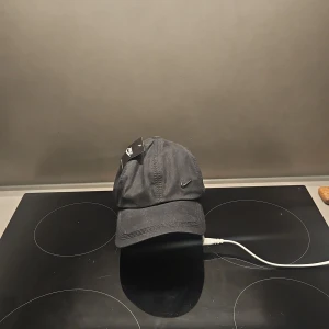 Svart keps från Nike - Snygg svart keps från Nike med klassisk böjd skärm och justerbar rem baktill. Kepsen har en liten broderad Nike Swoosh-logga på sidan och är tillverkad i bomull. Perfekt accessoar för en sportig och avslappnad stil.