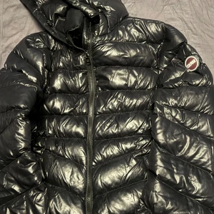 Svart pufferjacka från Colmar - Säljer en svart pufferjacka från Colmar med glansig finish och huva. Jackan har ett quiltat mönster, dragkedja framtill och Colmar-logga på ärmen. Perfekt för dig som vill ha en snygg och varm jacka med street vibe.