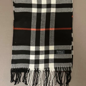 Rutig halsduk från Burberry - Snygg halsduk från Burberry med klassiskt rutigt mönster i svart, vitt och röda detaljer. Halsduken har fransar i ändarna och en diskret Burberrys-logga. Perfekt accessoar för att lyfta din stil under kyliga dagar. halsduken skickad från Norge och moms kan tilkomma. betalning måste ske via paypal eller liknande