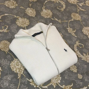 Vit stickad kofta från Polo Ralph Lauren - Vit ribbstickad kofta från Polo Ralph Lauren med hel dragkedja och klassisk krage. Tröjan har det ikoniska blå broderade logotypen på bröstet och är tillverkad i mjuk bomull. Perfekt för lager-på-lager och stilren look.