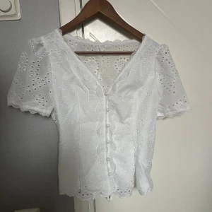 Vit spetsblus med knyt och knappar - Superfin vit blus med broderade hålmönster och spetsdetaljer längs v-ringningen och ärmsluten. Blusen har korta puffärmar, knappar framtill och ett knytband i midjan för extra form. 