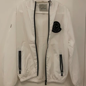 Vit vindjacka från Moncler med luva - Säljer en clean vit vindjacka från Moncler i storlek S. Jackan har svart dragkedja, två fickor med svarta detaljer, elastisk nederkant och luva. Moncler-logga på bröstet och armen ger extra premiumkänsla. Perfekt för dig som gillar stilrena och sportiga vibes.