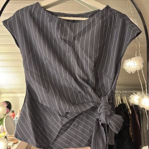 Randig omlottopp med knyt i midjan - Snygg mörkgrå omlottopp med vita ränder och knyt i midjan. Toppen har korta ärmar och en rak halsringning. Materialet känns lätt och mjukt, inte använd någon gång