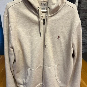 Beige hoodie från Polo Ralph Lauren - Snygg beige hoodie från Polo Ralph Lauren med dragkedja och snörning i huvan. Tröjan har två fickor framtill och det klassiska broderade logot på bröstet. Perfekt för chill dagar och enkel att matcha med jeans eller joggers.