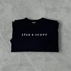 Svart Lyle & Scott t-shirt med tryck - Svart t-shirt från Lyle & Scott med klassiskt vitt tryck på bröstet. Modellen är rak med rund hals och korta ärmar. Materialet är mjuk bomull som känns skönt mot huden. Perfekt för dig som gillar stilrena och enkla plagg med tydlig logga.