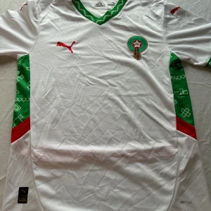 Marocko vit fotbollströja Puma - Säljer en officiell Marocko fotbollströja  i vitt med gröna och röda detaljer. Tröjan har korta ärmar, v-ringad krage med grönt mönster och Marockos landslagsemblem på bröstet. Tillverkad i lätt och ventilerande polyester med DryCell-teknologi.
