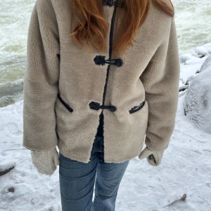 Beige teddyjacka från mango - Säljer en beige teddyjacka med coola svarta snörningsdetaljer framtill och vid fickorna. Jackan har rund hals, lång ärm och loose passform. Materialet är mjukt och fluffigt, perfekt för kalla dagar. Enkel och stilren design som är lätt att matcha med jeans eller andra plagg.