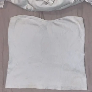 Vit bandeau tubtopp från Gina Tricot - Säljer en vit bandeau tubtopp från Gina Tricot i storlek M. Toppen är ribbad och har en mjuk, elastisk passform som sitter snyggt runt kroppen. Perfekt för att styla med jeans eller kjol. Enkel och clean design utan ärmar.