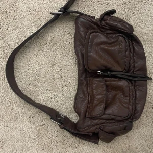 Brun axelväska i skinn från urban outfitters - Säljer en brun axelväska från urban outfitters i mjukt skinn med flera praktiska fickor och justerbar rem. Väskan har coola dragkedjedetaljer och en snygg, lite slouchy form som ger en chill vibe. Perfekt storlek för det viktigaste och passar till många olika outfits.