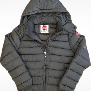 Colmar jacka - Snygg grå pufferjacka från Colmar Originals med huva och dragkedja framtill. Jackan har ett quiltat mönster och elastiska muddar vid ärmslut. Märkets logga i rött och vitt syns på ärmen och insidan. Perfekt för kalla dagar. Lappen med storlek nere i jackan är borta (försvann när jag tvättade jackan)