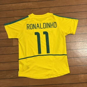 Brasilien Ronaldinho fotbollströja Nike - Gul Brasilien fotbollströja från Nike med gröna detaljer och nummer 11, samt Ronaldinho tryckt på ryggen. Tröjan har korta ärmar, rund hals och meshpaneler för extra ventilation. Perfekt för dig som älskar klassisk fotbollsstil och ikoniska spelare.