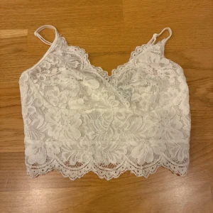 Vit spetsig croppad linnetopp - Säljer en vit croppad linnetopp med blommig spets och tunna axelband. Toppen har v-ringad hals och en snygg dragkedja i ryggen. Spetsen har fina detaljer och vågig kant nedtill som ger en extra touch. Perfekt för dig som gillar romantisk stil.