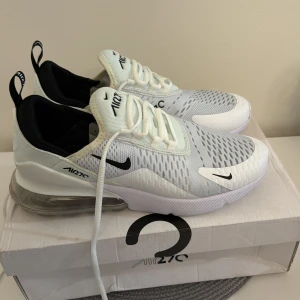 Nike Air Max 270 vita sneakers - Nike Air Max 270 sneakers i vit och svart med mesh-överdel och synlig Air Max-sula. Skorna har snörning, rund tå och platt sula. Snygg design med svarta detaljer och logga på både plös och häl. Perfekta för dig som gillar sportig och modern stil.