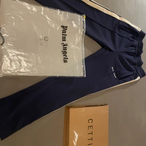 Palm Angels blå trackpants med vita ränder - Snygga marinblå trackpants från Palm Angels med vita sidoränder och broderad logga på benet. Byxorna har elastisk midja och raka ben, tillverkade i polyester för en sportig vibe. Perfekta för dig som gillar streetwear och vill sticka ut. Kvitto medföljer digitalt, skriv för mera funderingar.