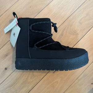 Svarta vinterskor från Tretorn - ”Moon boots” från Tretorn i svart med grov platt sula och snörning framtill. Utsidan är i syntet och mocka, insidan är fodrad med mjuk päls för extra värme. Skorna har rund tå och detaljerade snören med reflextråd. Helt nya. Kommer med låda och tags.
