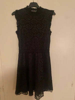 Svart spetsklänning med hög hals, 13-14 år - Svart klänning i spets med hög hals och ärmlös design från Cubus. Resår i midjan och lätt utställd kjol. Storlek 13-14 år.