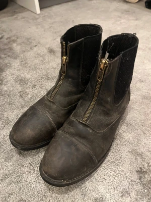 Svarta ridstövletter från CRW - Säljer ett par svarta ridstövletter från CRW i skinn med dragkedja framtill och elastiska paneler på sidorna. Skorna har rund tå och platt sula, perfekt för dig som gillar ridning och vill ha en klassisk look. Snygga detaljer med guldiga dragkedjor.