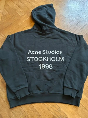 Svart hoodie Acne Studios Stockholm - Bra skick! Svart hoodie från Acne Studios med stor vit text på ryggen: 'Acne Studios STOCKHOLM 1996'. Klassisk modell med huva och ribbade muddar. Mjuk och skön, perfekt för chill dagar. Oversized passform för extra comfy vibe. Skriv om du har frågor!