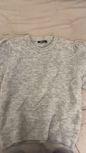 Grå stickad tröja från Gina Tricot - Mysig grå stickad tröja från Gina Tricot med korta puffärmar och rund hals. Tröjan har ribbade muddar vid ärmslut och nederkant, vilket ger en snygg och enkel look. Perfekt för dig som gillar stilrena och bekväma plagg.