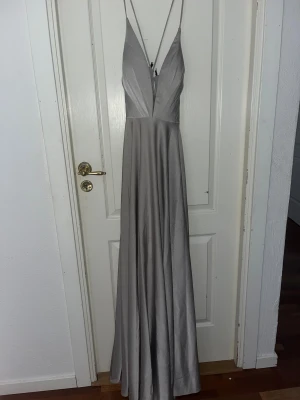 Silvrig balklänning med snörning - Säljer en elegant silver färgad balklänning med djup v-ringning och smala axelband. Klänningen har snygg snörning i ryggen, dragkedja i ryggen och praktiska fickor. Materialet är glansigt och känns lyxigt, perfekt för dig som vill känna dig som en prinsessa på balen. Nypris 3000kr (liten fläck på framsidan som syns på sista bilden, skulle inte säga det syns så mycket i verkligheten däremot)