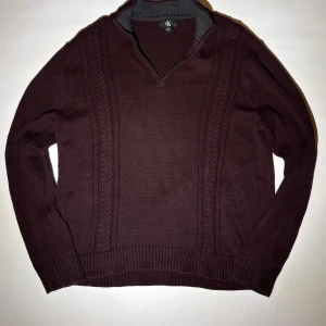 Vinröd stickad tröja med half zip - Snygg vinröd stickad tröja från GK med half zip och ribbade muddar. Tröjan har ett diskret kabelstickat mönster framtill och en hög krage med grå insida. Perfekt för dig som gillar klassisk stil med en modern twist.