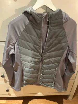 Grå quiltad pufferjacka från OTP - Snygg grå pufferjacka från OTP med quiltad framsida och rygg, huva och hel dragkedja. Jackan har långärmade släta ärmar och två sidofickor med dragkedja. Perfekt för dig som gillar sportig och clean stil. Liten i storleken.