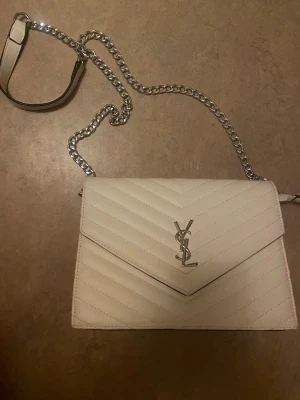 Vit quiltad axelväska från YSL - Snygg vit axelväska från YSL med silverkedja och ikonisk YSL-logga framtill. Perfekt storlek för att bära med sig det viktigaste och ger en lyxig vibe till din outfit.