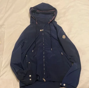 Mörkblå vindjacka från Moncler - Snygg mörkblå vindjacka från Moncler med huva och dragkedja framtill. Jackan har snörning vid huvan, silverfärgade detaljer och Moncler-logga på ärmen. Tillverkad i lätt polyester, perfekt för blåsiga dagar. Fickor med dragkedja och stilren design. Den har ett liten hål som man kan se på näst sista bilden men man ser inte de så mkt.