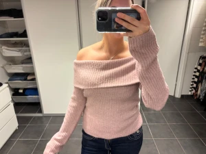 Ljusrosa ribbad offshoulder stickad tröja - Superfin ljusrosa stickad tröja med ribbad struktur och offshoulder-design. Tröjan har långa ärmar och sitter snyggt med en lite tight passform. Perfekt för dig som vill ha en trendig och mysig look med lite extra edge.