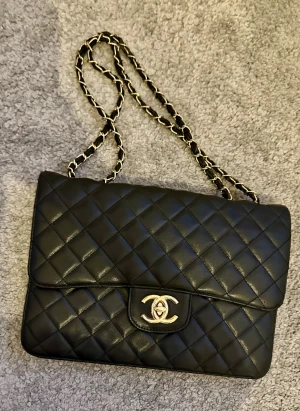 Snygg / Svart axelväska - Säljer en svart axelväska i skinn med quiltat mönster och klassisk CCc-logga i metall framtill. Väskan har en kedja med invävt skinn och röd insida med dragkedja. Perfekt storlek för det viktigaste och riktigt snygg att bära över axeln.