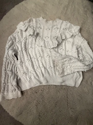 Vit stickad tröja med volang från H&M - Supersöt vit stickad tröja från H&M med volangdetaljer över axlar och ärmslut. Tröjan har ett snyggt kabelstickat mönster med små bollar och ribbad hals. Perfekt för dig som gillar romantisk och trendig stil.