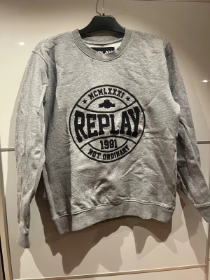 Grå sweatshirt från Replay - Snygg grå sweatshirt från Replay med stort svart broderat logotryck på bröstet. Rund halsringning och långa ärmar med ribbade muddar. Mjuk och skön bomullsmix som passar perfekt för chill dagar. Klassisk passform och cool streetstil.