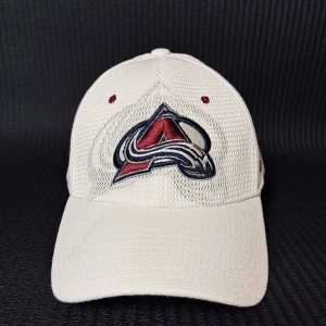 Vit Colorado Avalanche keps Zephyr - Snygg vit keps från Zephyr med Colorado Avalanche-logga broderad fram och bak. Kepsen har meshstruktur, röda detaljer och böjd skärm. NHL-märkt och officiell merchandise, perfekt för dig som älskar hockey och vill representera ditt lag.