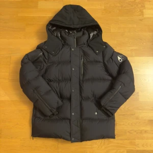 Svart pufferjacka från Moose Knuckles - Säljer en svart pufferjacka från Moose Knuckles med ikonisk logga på ärmen och insidan. Jackan har huva, tvåvägsdragkedja, tryckknappar och flera fickor med dragkedja. Materialet är polyester och dun, perfekt för kalla dagar. Snygg och stilren design med mörk ton.          Pris kan diskuteras, den är äkta och kvitto finns!